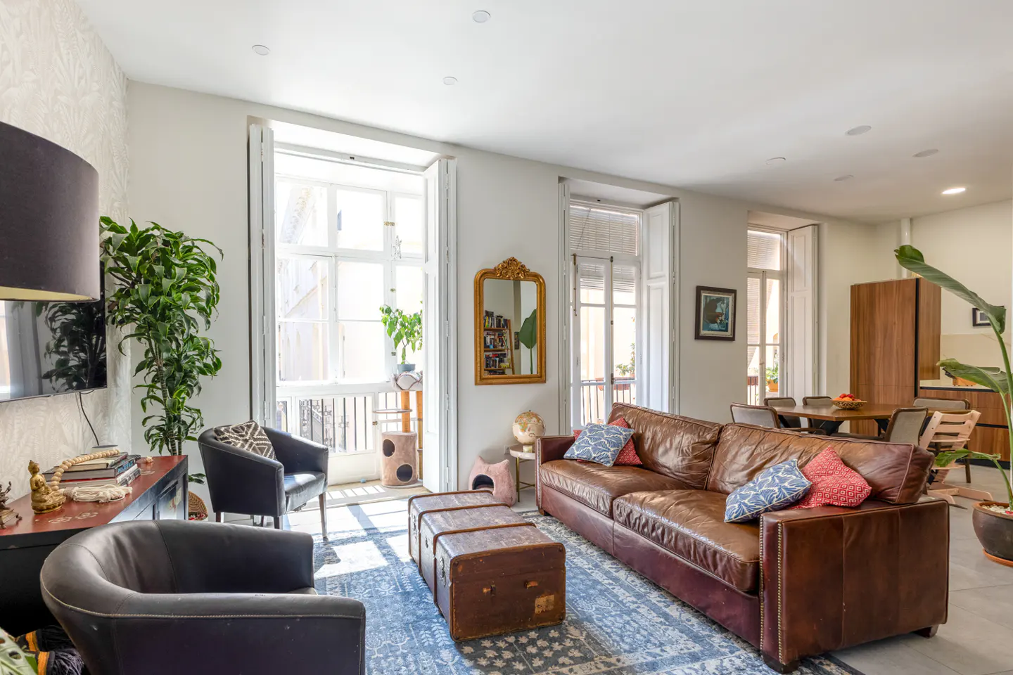 Bright living room with a brown leather sofa, trunk coffee table, and blue patterned rug. Balcony access through white-framed doors.