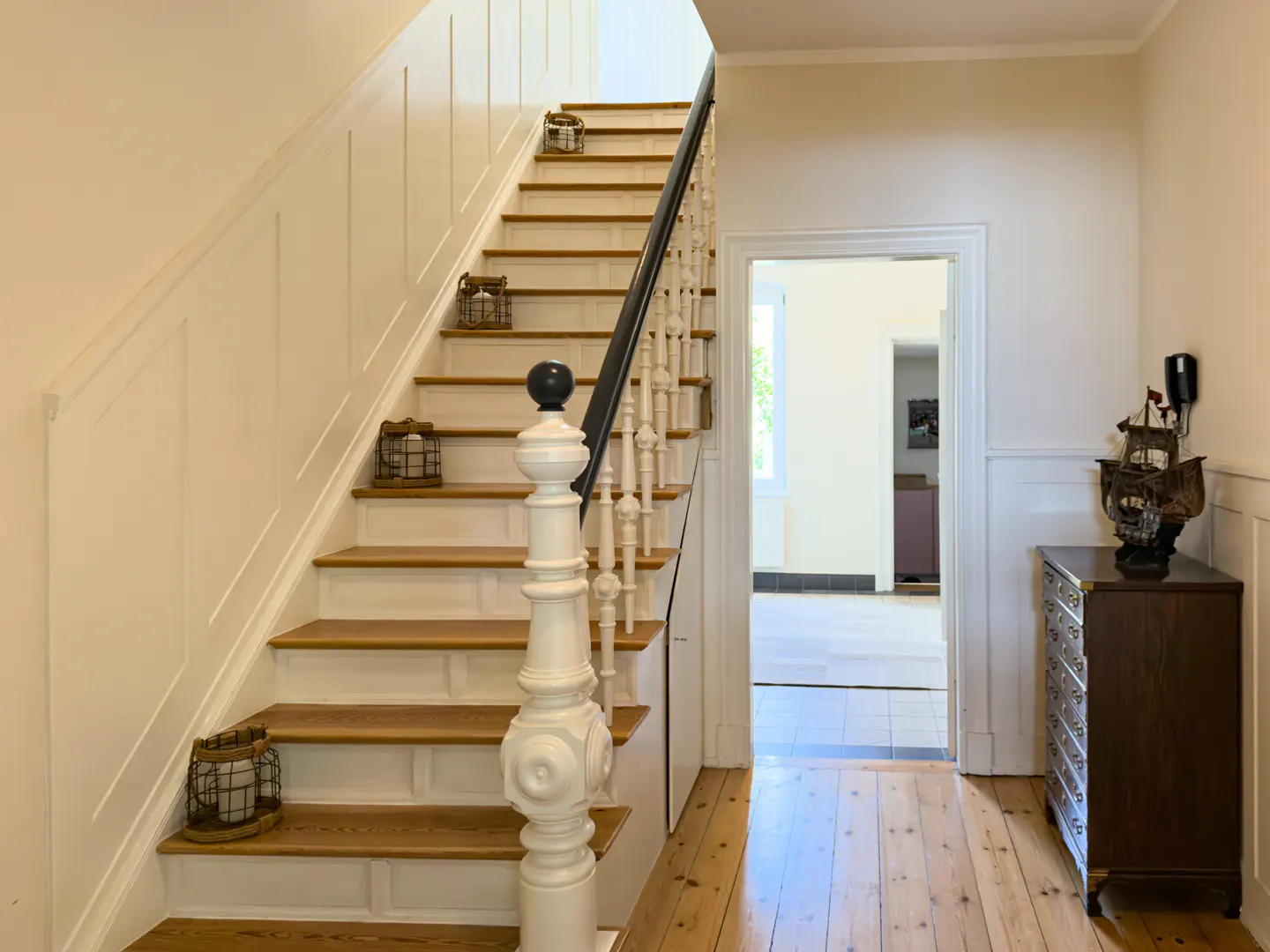Interior view of a hallway with a staircase, wooden floors, and a dark wood dresser with a ship model on top.