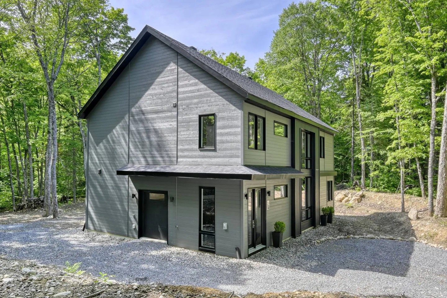 Two-story gray house with black trim and a black roof, surrounded by green trees and a gravel driveway.
