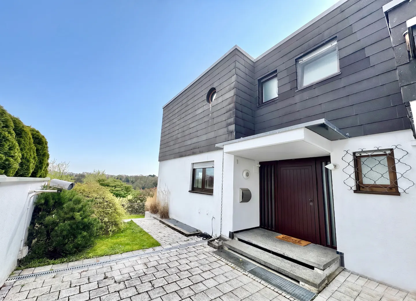 Modern two-story house with gray siding and a dark brown front door. White walls, stone driveway, and green landscaping. Blue sky.