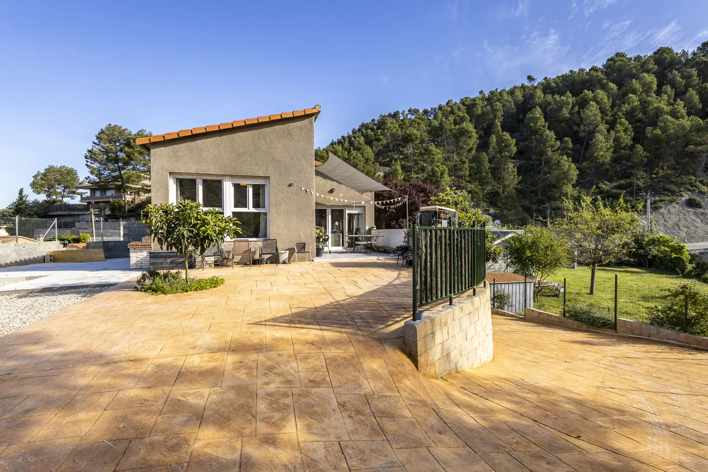 Exterior view of a modern, single-story home with a beige facade and orange roof tiles, surrounded by a large, patterned patio and lush greenery.