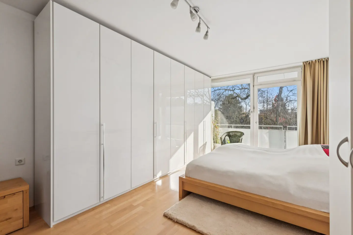 Bright bedroom with a wooden bed frame, white bedding, and a wall of white closets. A balcony is visible through the glass doors.