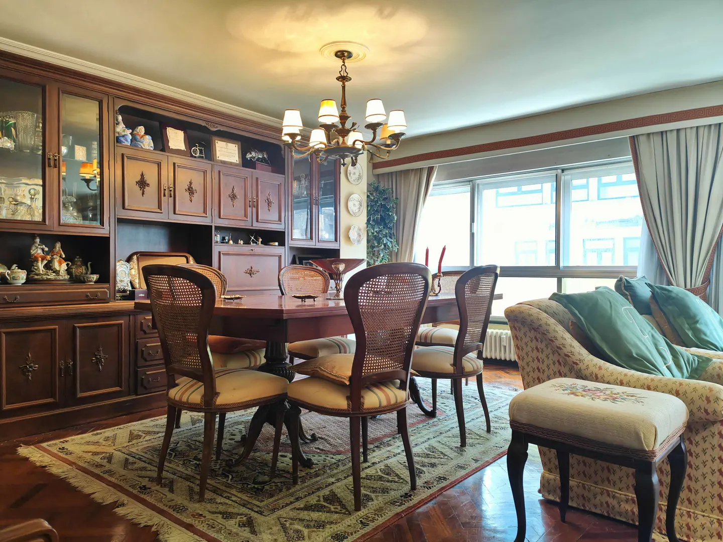Dining room with a wooden table, cane-back chairs, large hutch, chandelier, and a sofa with green pillows on a rug.