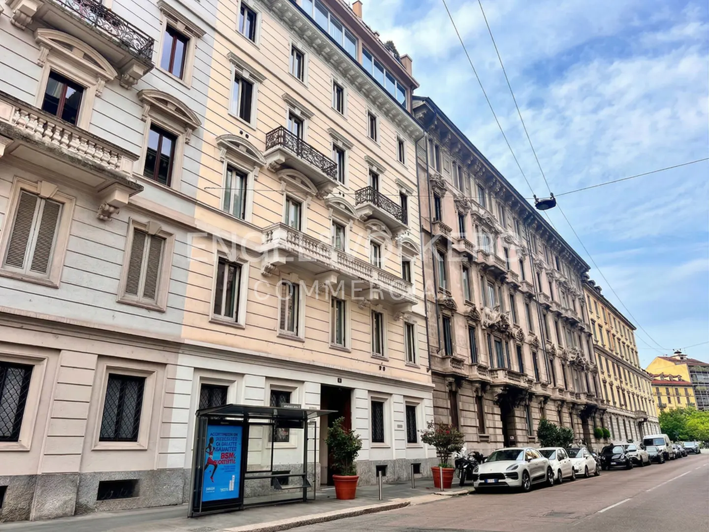 Street view of multi-story buildings with balconies, a bus stop, and parked cars on a sunny day.