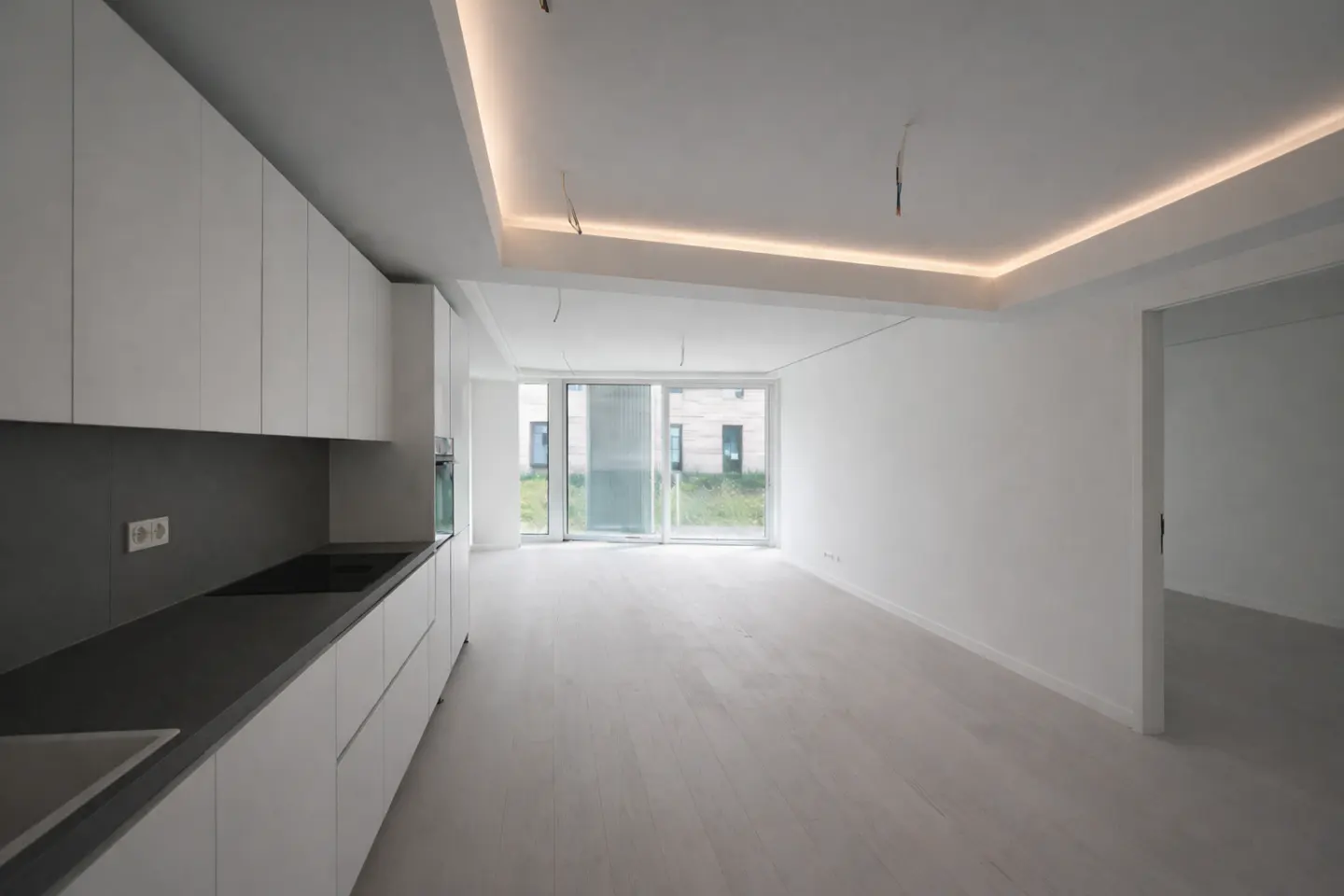 A bright, modern apartment interior with white walls, cabinets, and light wood floors. A sliding glass door leads to a view of a building.
