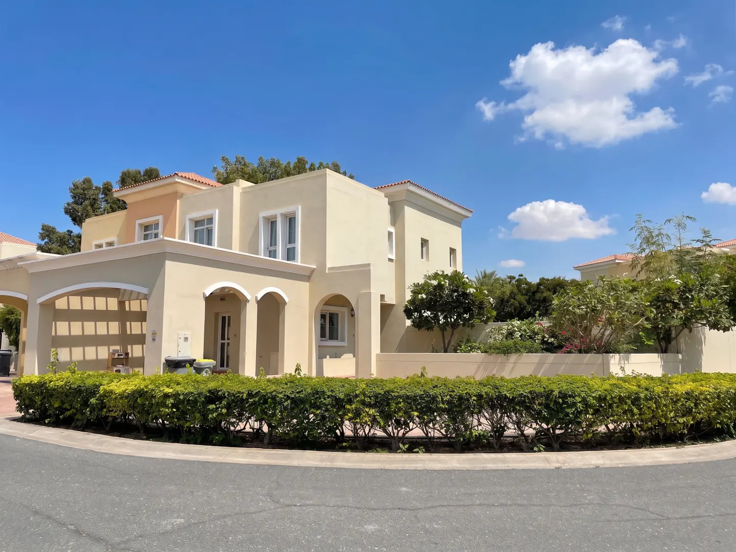 A two-story beige house with a red tile roof under a blue sky with white clouds. Green bushes line the front of the property.