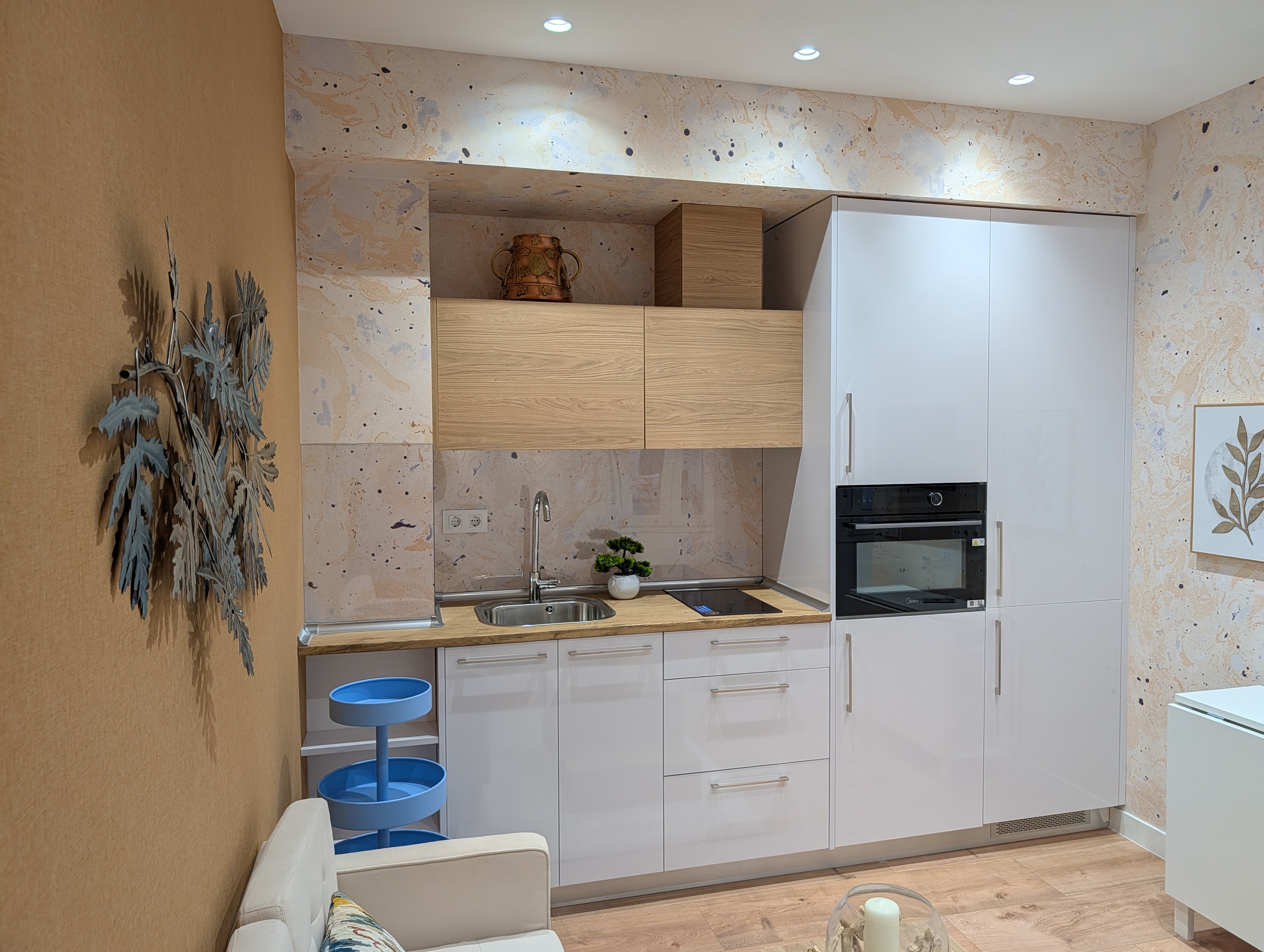 Modern kitchen with white cabinets, wood-tone upper cupboards, speckled backsplash, and a blue tiered cart in a studio space.