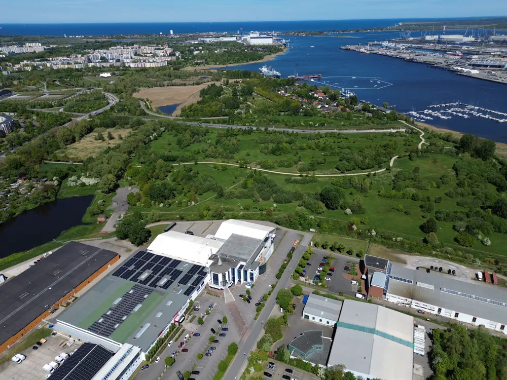 Aerial view of a commercial building with solar panels, surrounded by green spaces, a harbor, and a city skyline under a blue sky.