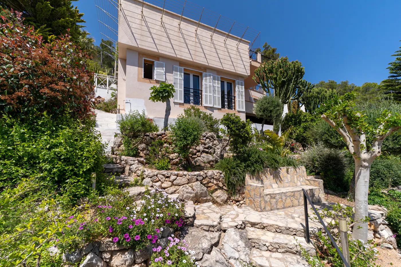 Exterior view of a two-story house with white shutters, surrounded by lush greenery and stone steps leading up to the entrance.