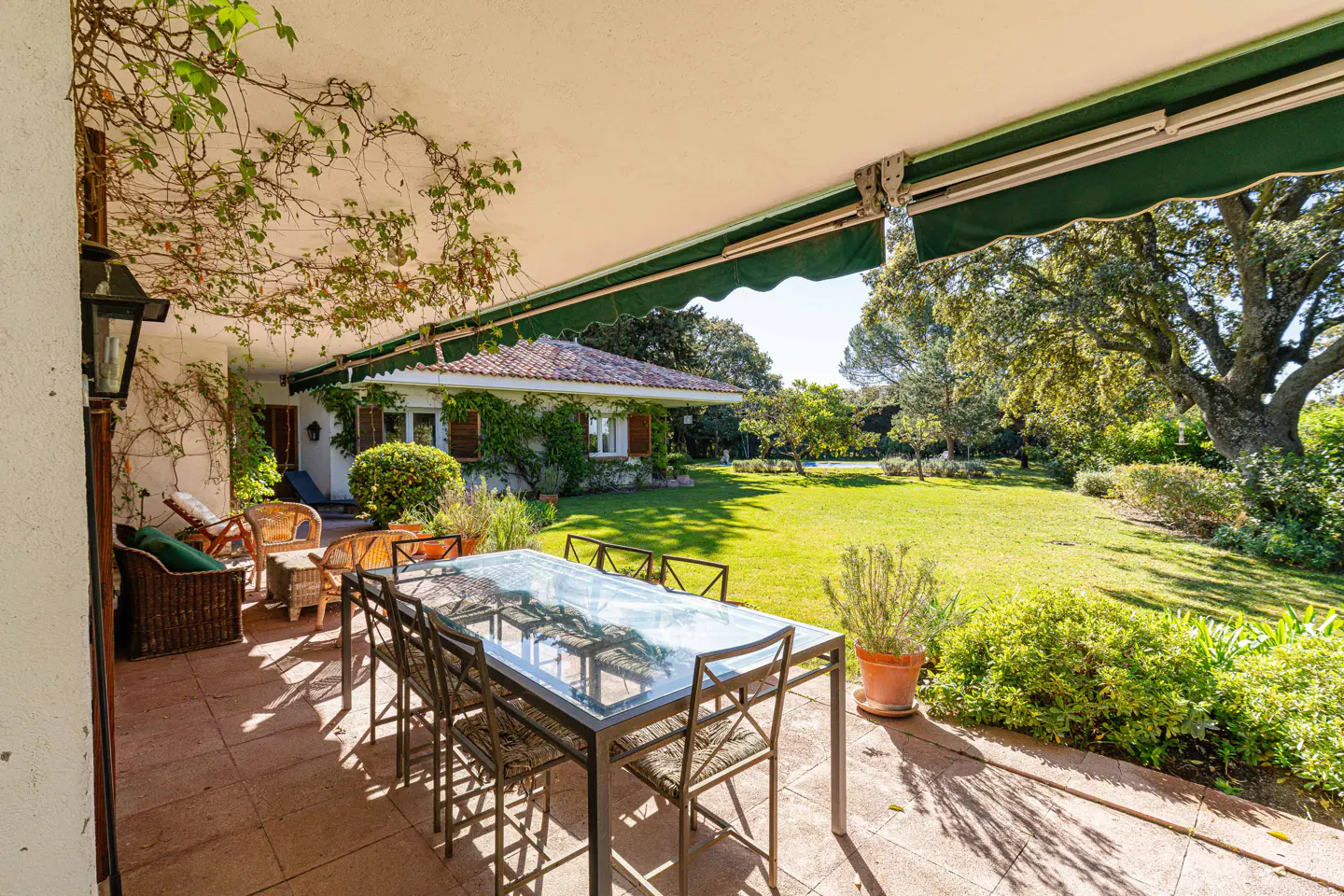 Covered patio with a glass table and chairs, overlooking a green lawn and trees. The house has a red tile roof and climbing vines.