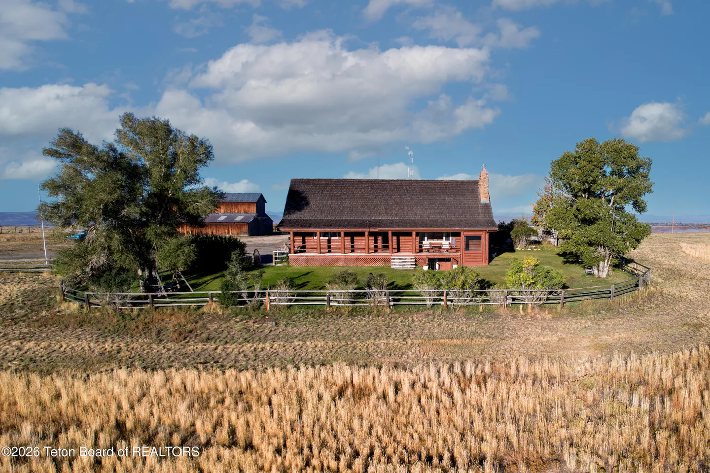 A red log cabin with a brown roof sits on a grassy hill surrounded by a wooden fence and tall grass under a blue sky.