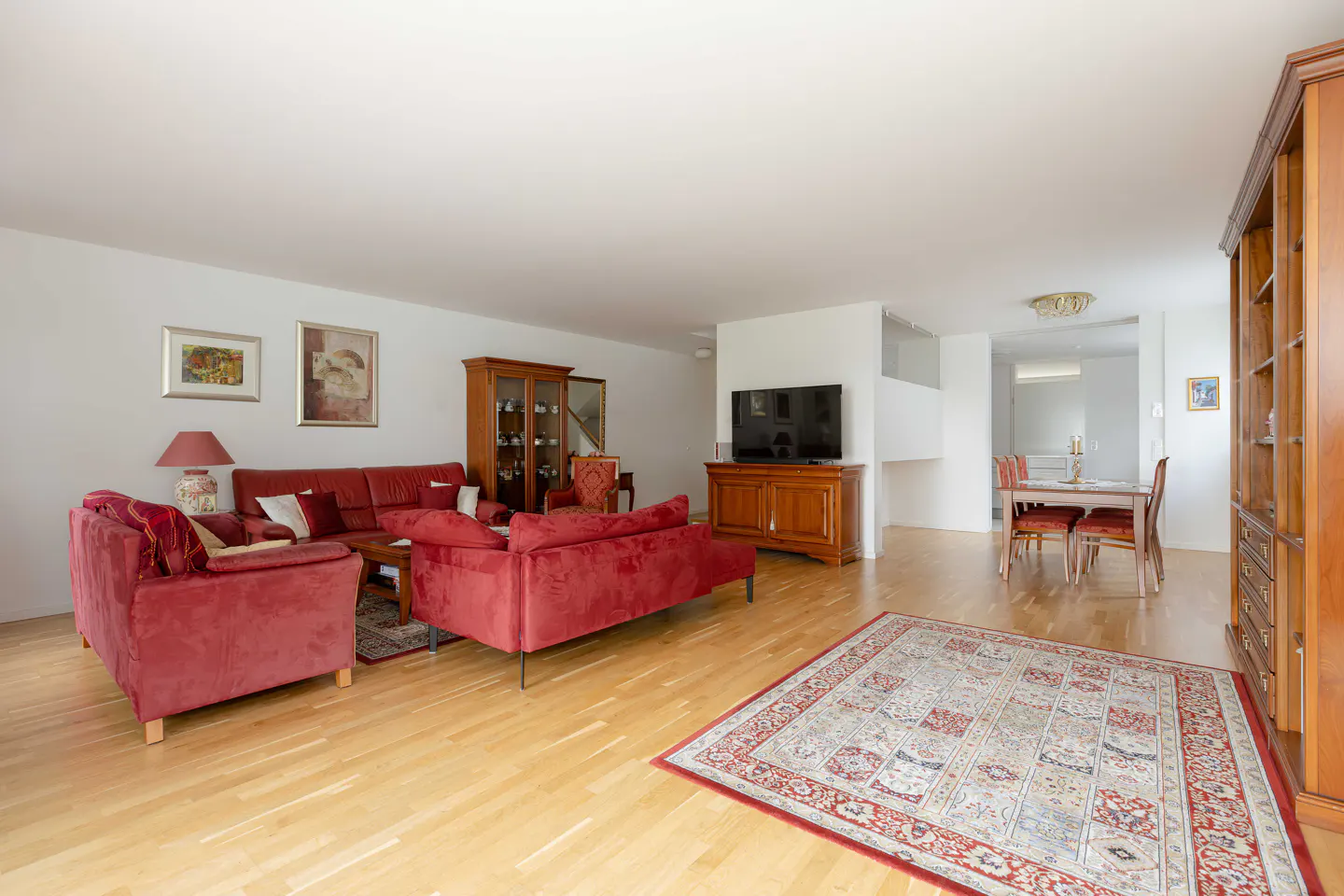 Bright living room with red sofas, wood floors, and a patterned rug. A dining table is visible in the background.