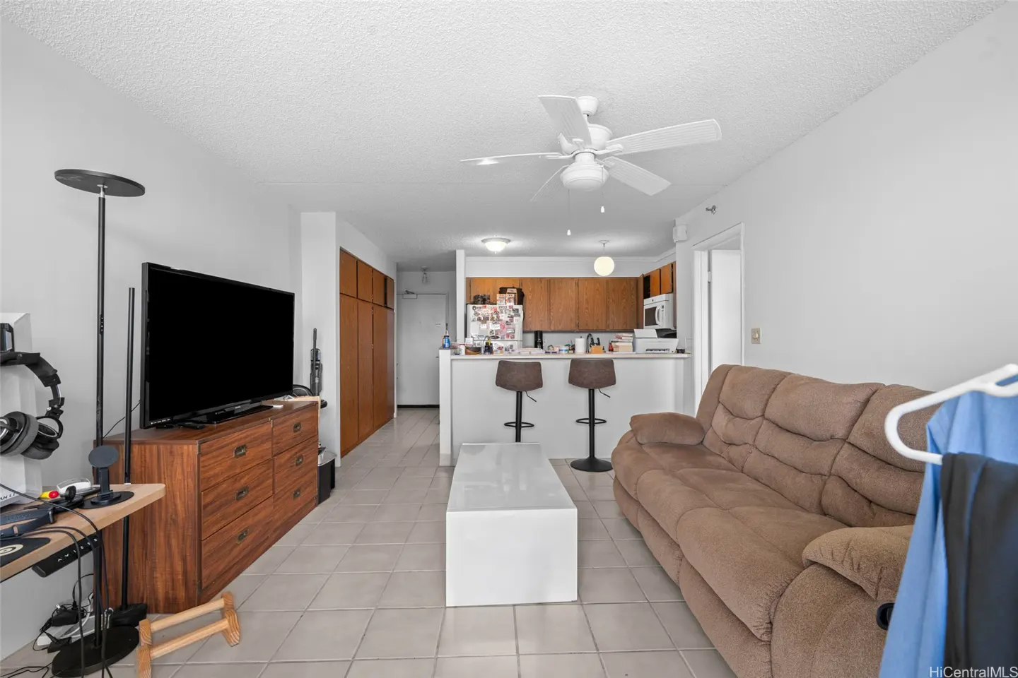 Living room with a brown sofa, white coffee table, TV on a wooden dresser, and a kitchen with bar stools in the background.