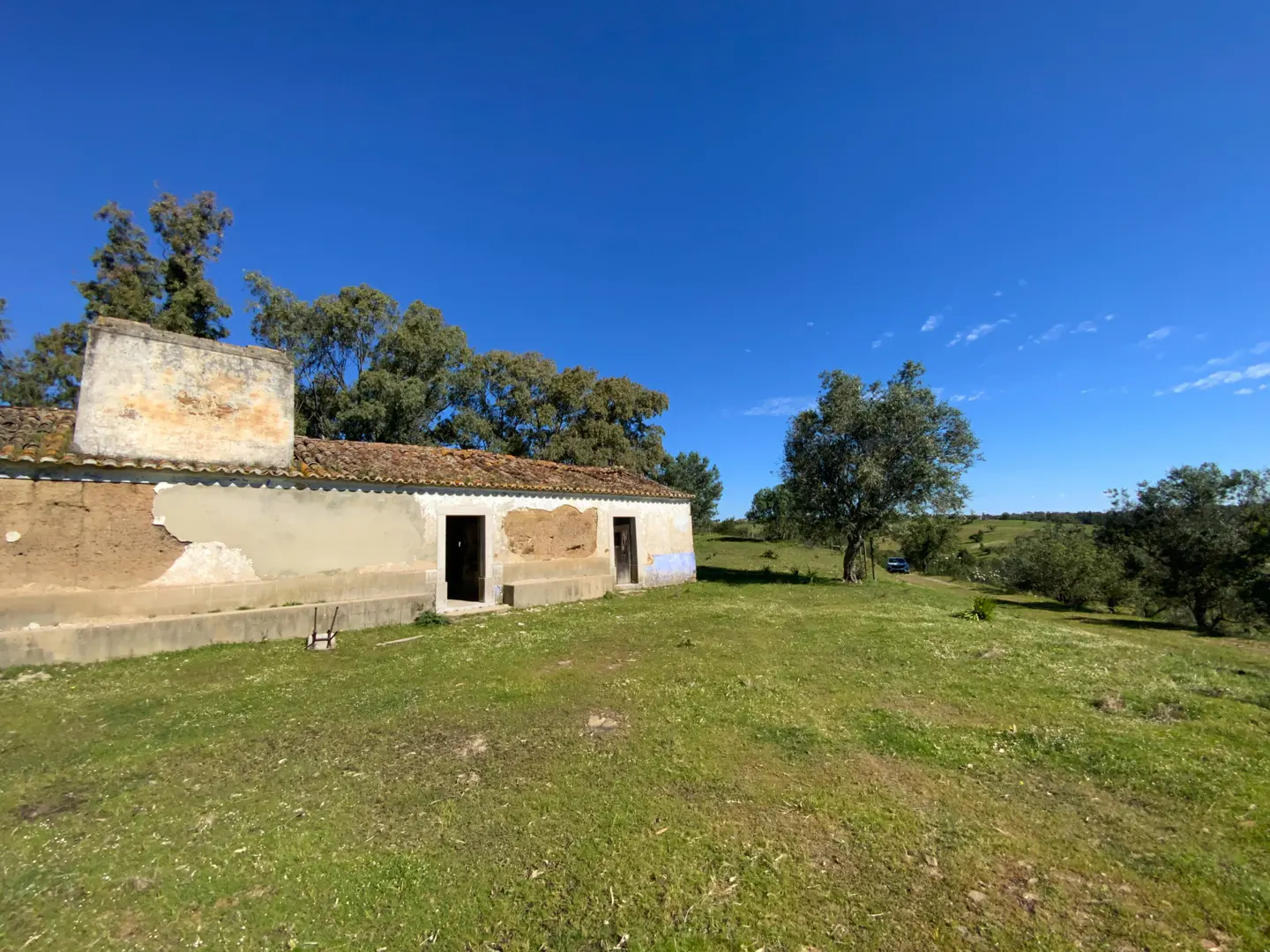 Exterior view of a dilapidated, one-story building with a red tile roof, set in a green field under a clear blue sky.
