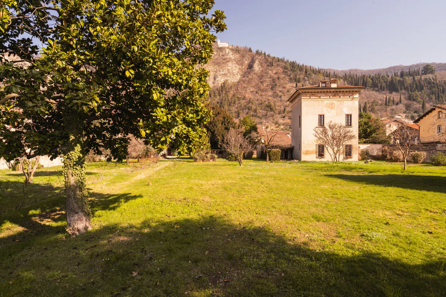 Exterior view of a light-colored, three-story building with a red tile roof, surrounded by a green lawn and trees. A mountain is in the background.