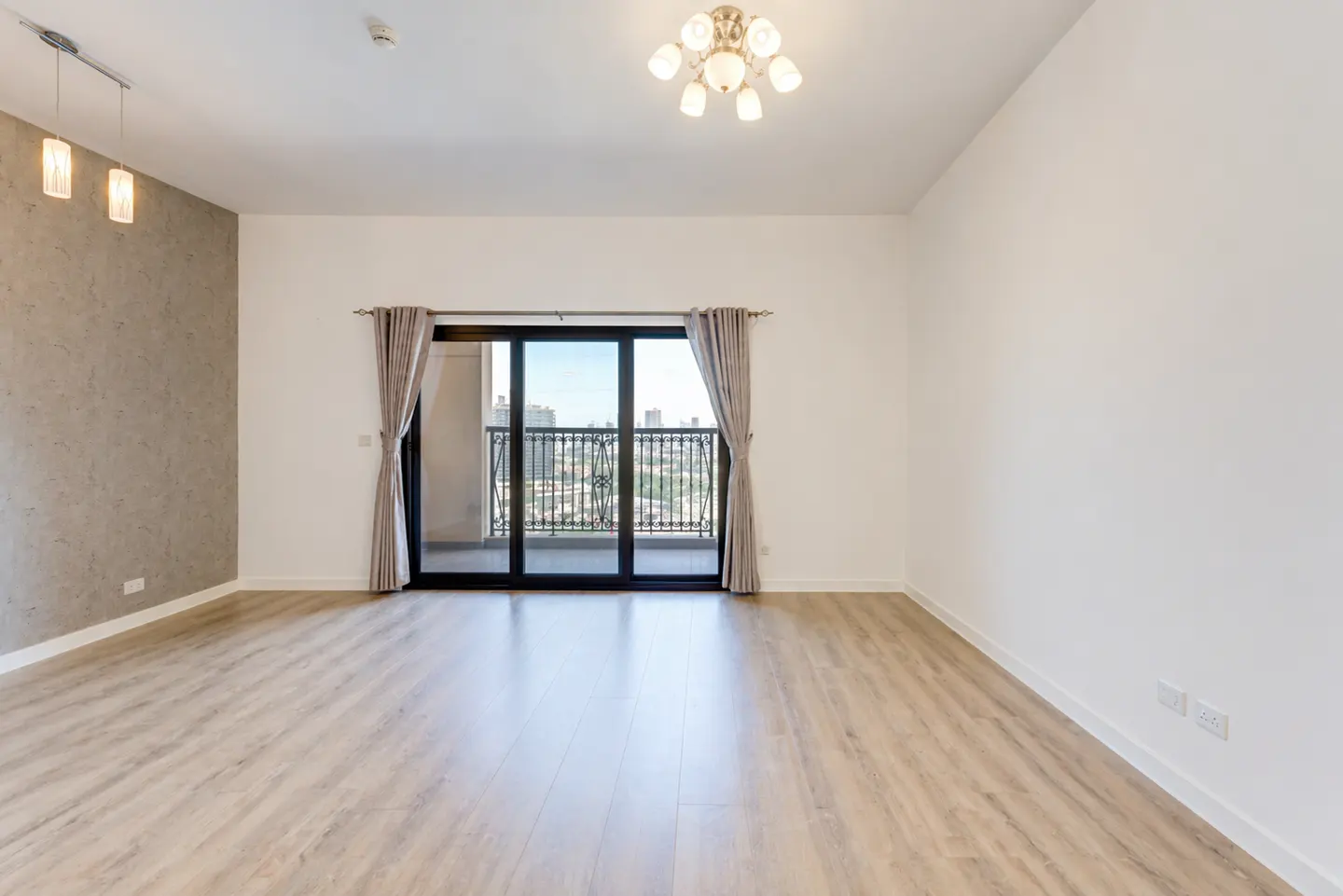 Empty living room with light wood floors, white walls, and a gray accent wall. Sliding glass doors lead to a balcony with city views.
