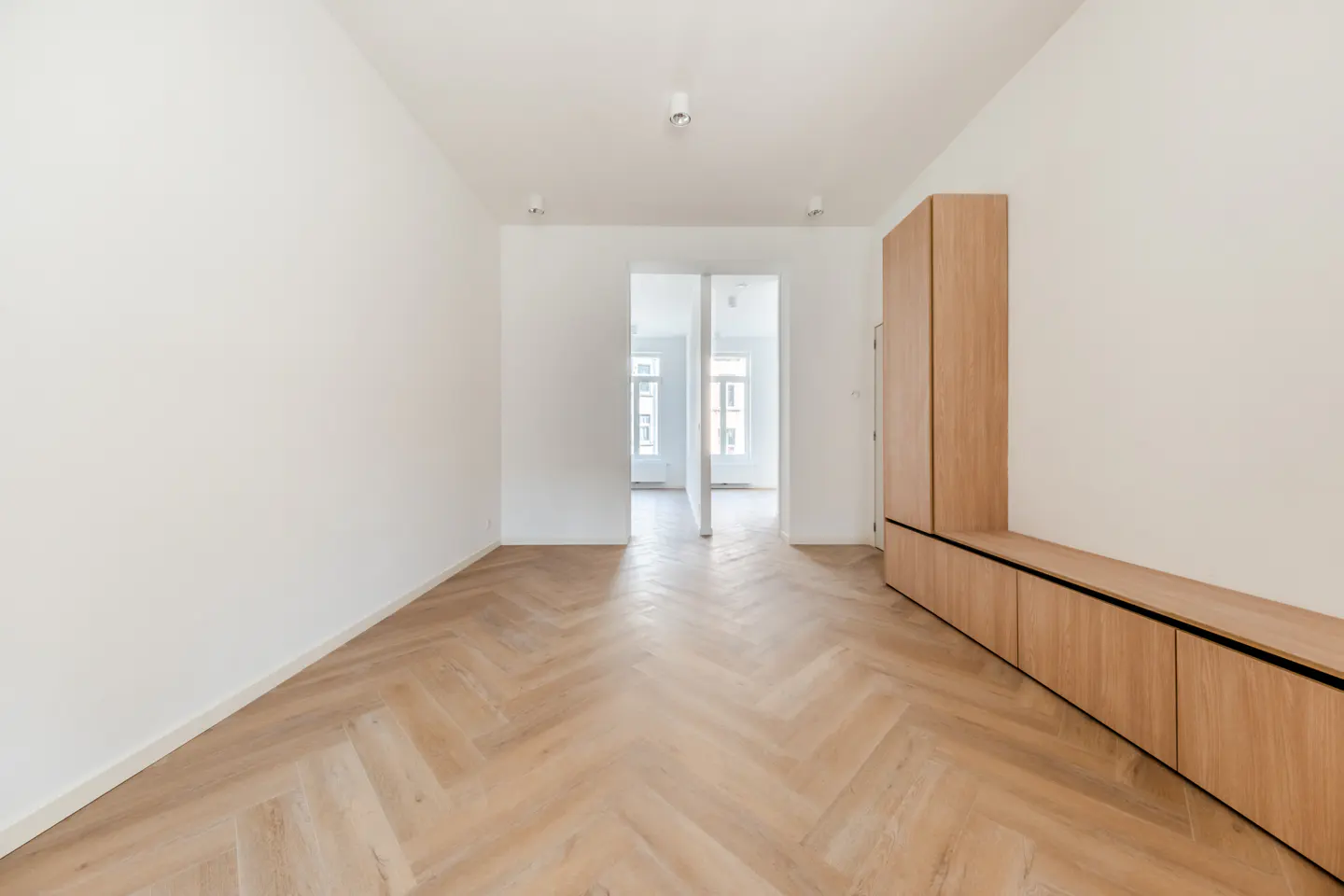 Bright, empty room with white walls, herringbone wood floor, and light wood cabinet. Doorway leads to another room with windows.