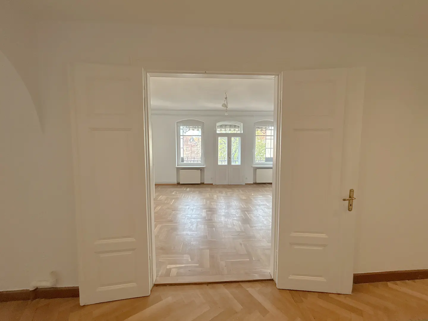 View through open white double doors into a bright, empty room with hardwood floors and three windows.