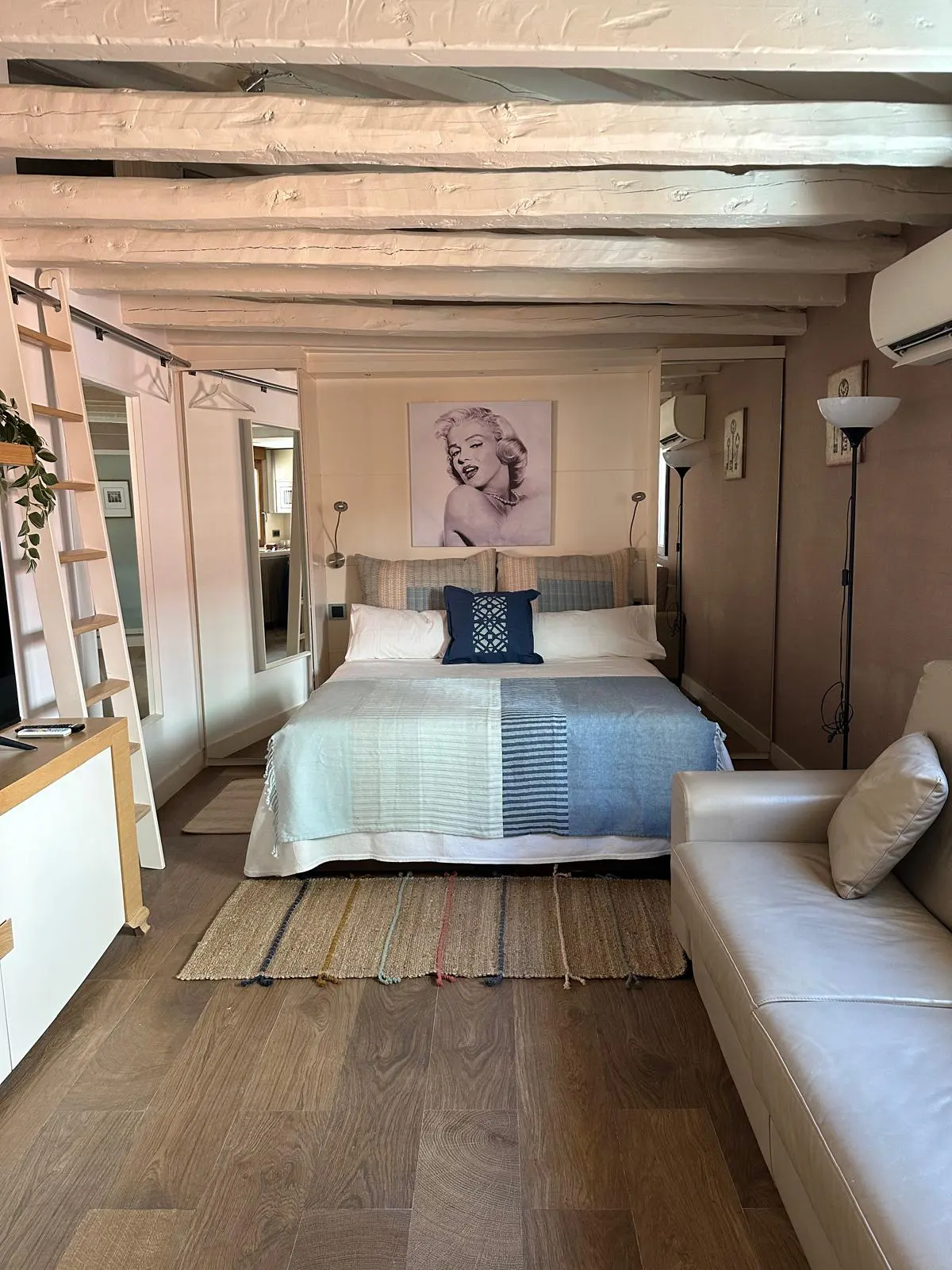 Bedroom with exposed white beams, a king bed with blue bedding, Marilyn Monroe art, and a white sofa on wood floors.
