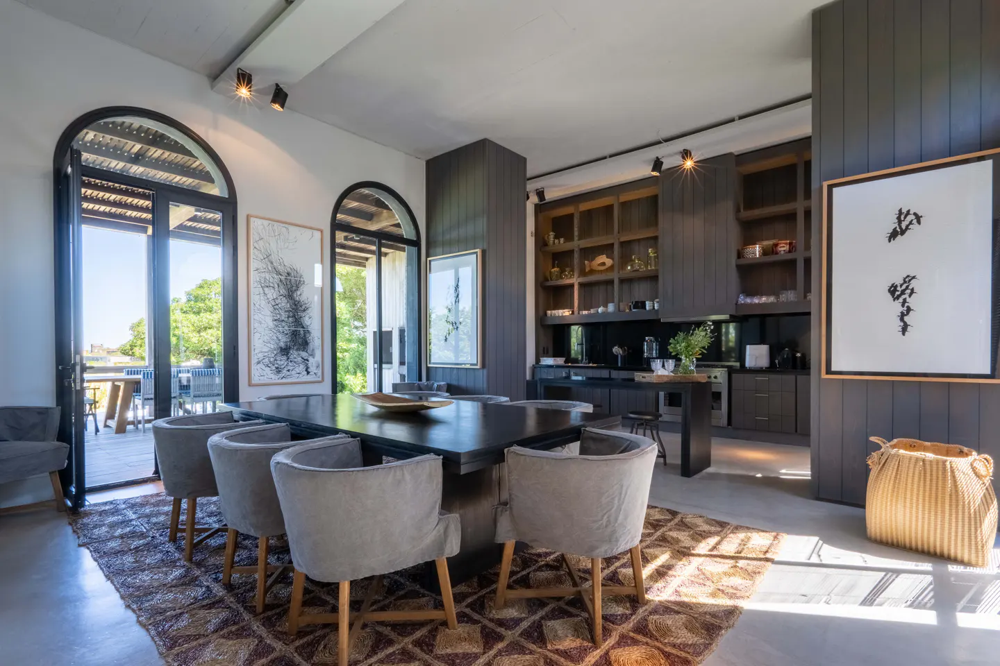 A modern dining room with a dark wood table, gray chairs, and a patterned rug. Arched windows lead to a patio. Dark wood cabinets and artwork adorn the walls.
