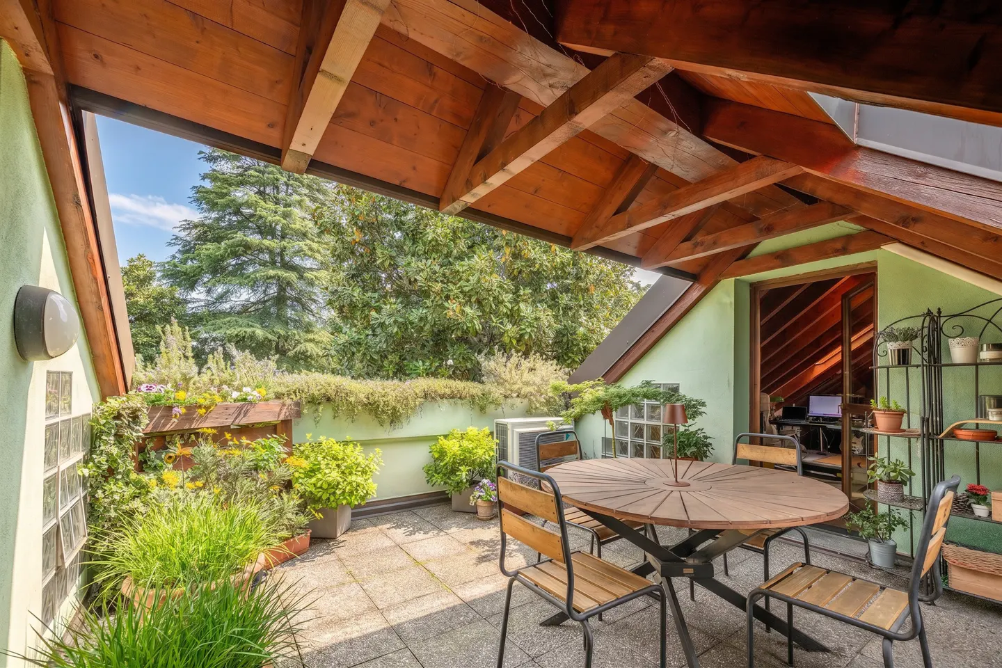 A cozy rooftop patio with a wooden table, chairs, and potted plants. The patio has a wooden roof and a view of lush green trees.