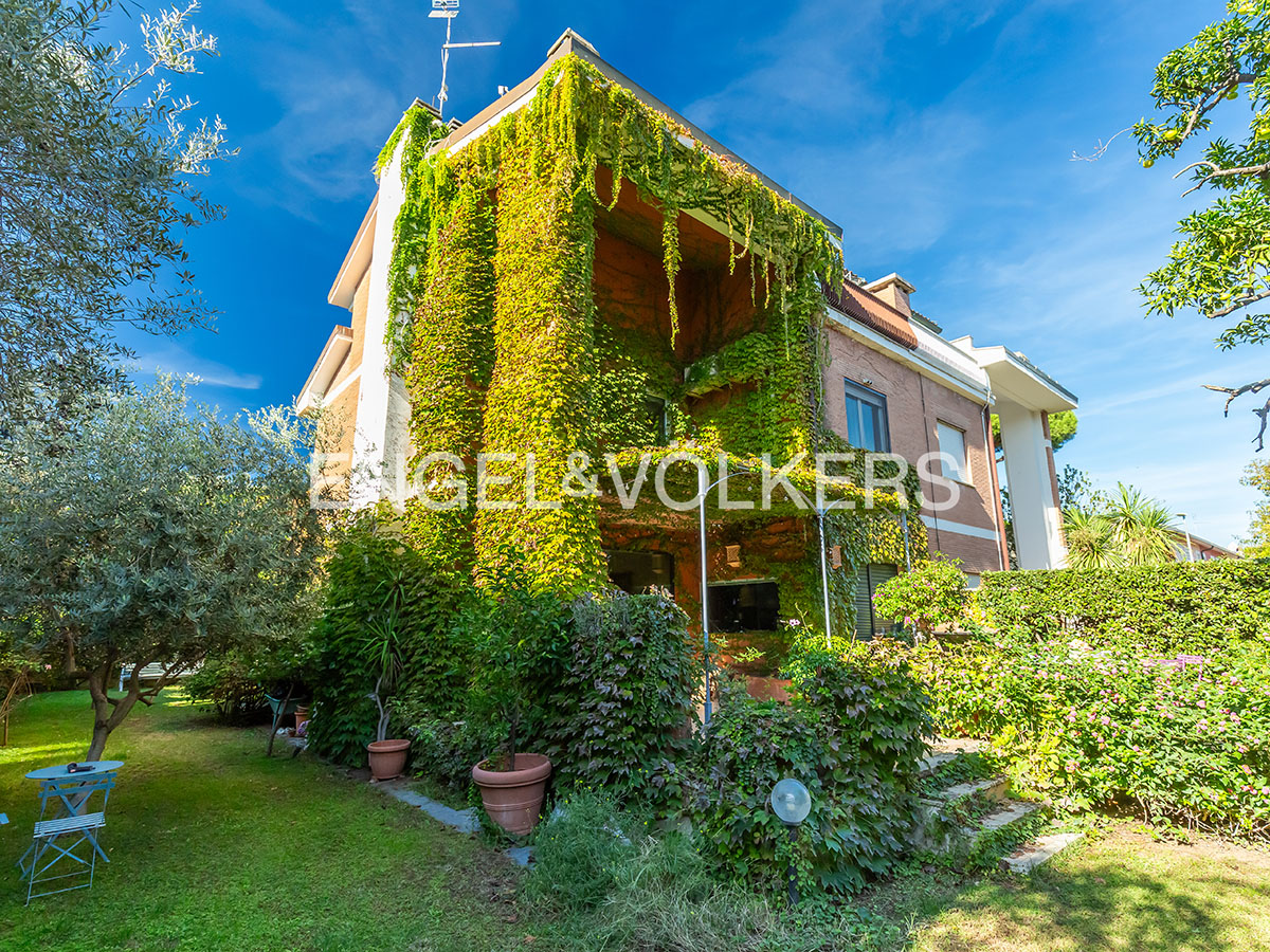 A multi-story house covered in lush green ivy, set in a sunny garden with trees, potted plants, and a small patio table. [AI-generated text]