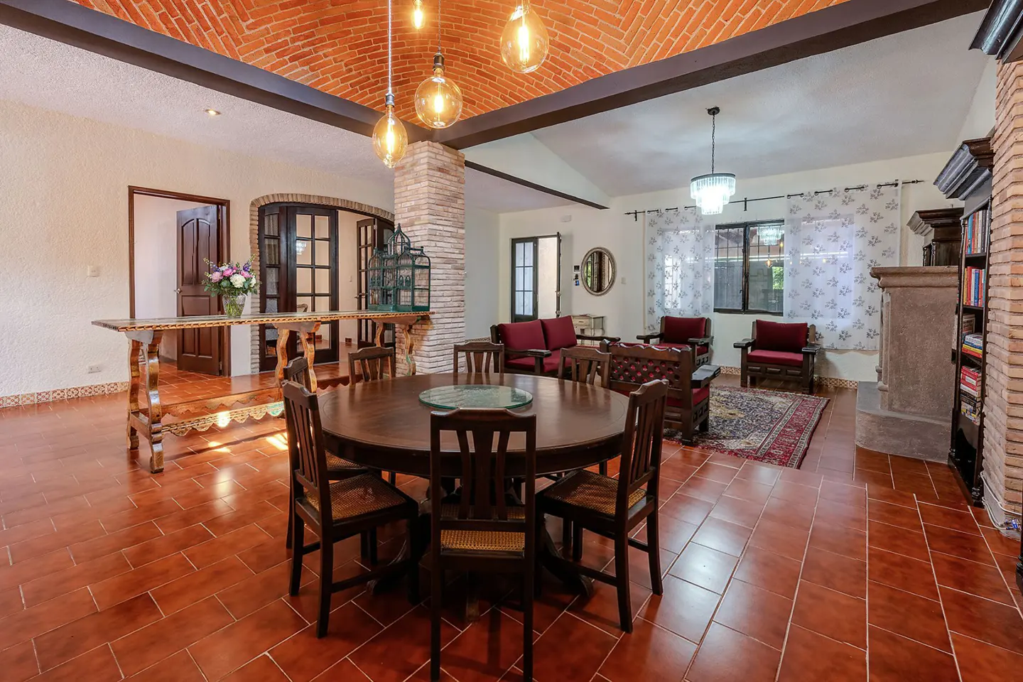A spacious living room with a brick ceiling, terracotta tile floor, and dark wood furniture. A round table with chairs is in the foreground.