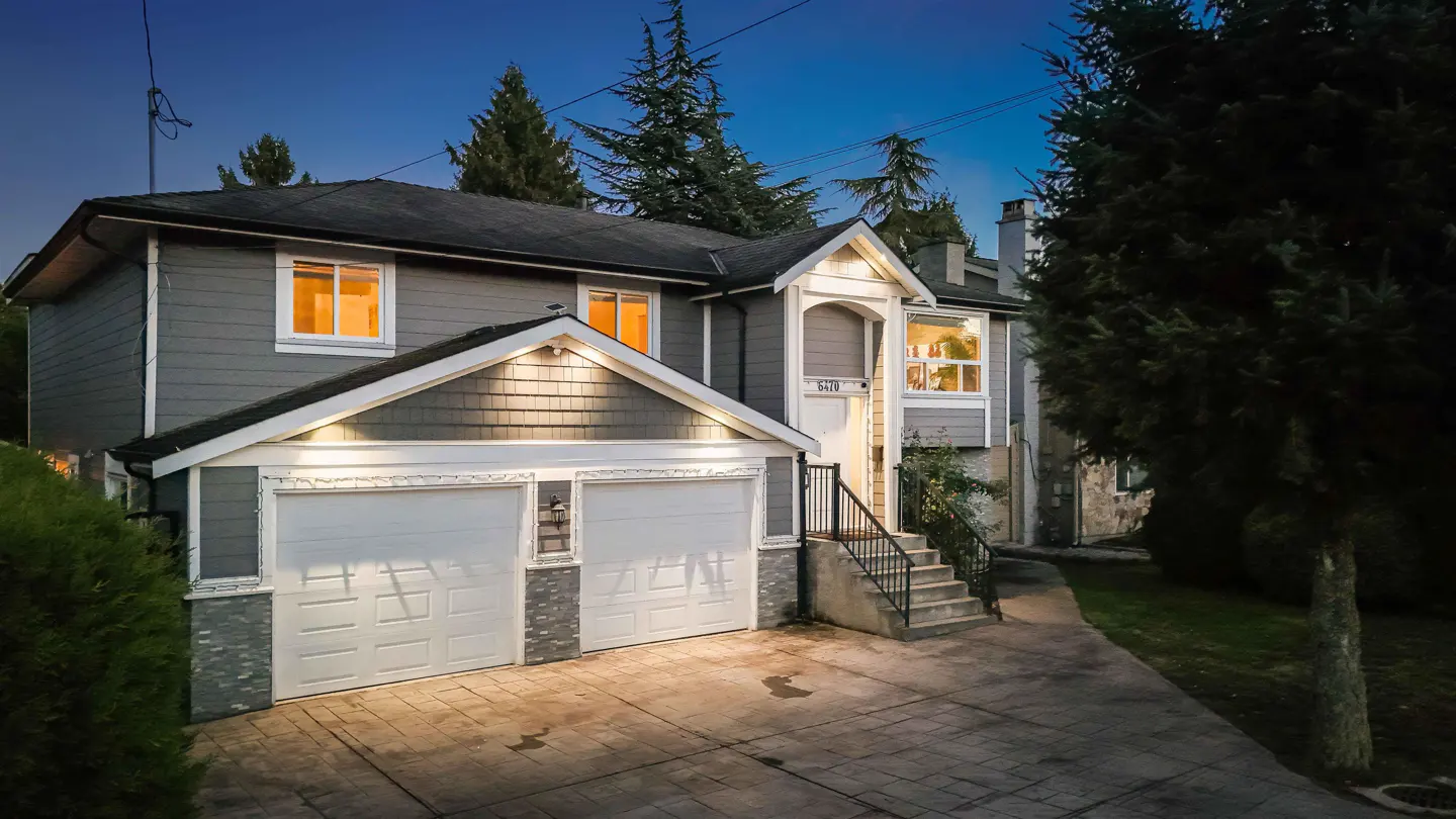 Two-story gray house with a two-car garage and a concrete driveway at dusk. The windows are lit, and trees surround the property.