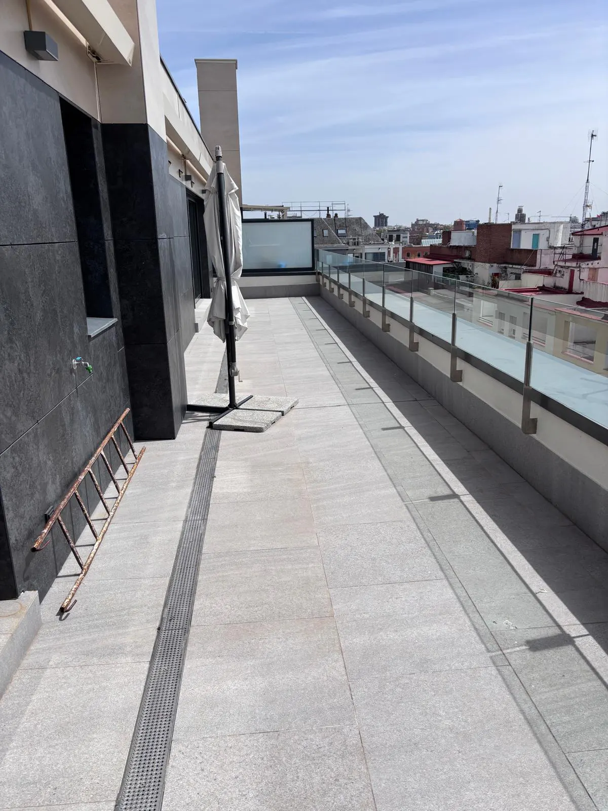 A rooftop terrace with gray stone tiles, a closed white umbrella, and a glass railing overlooking a cityscape under a blue sky.