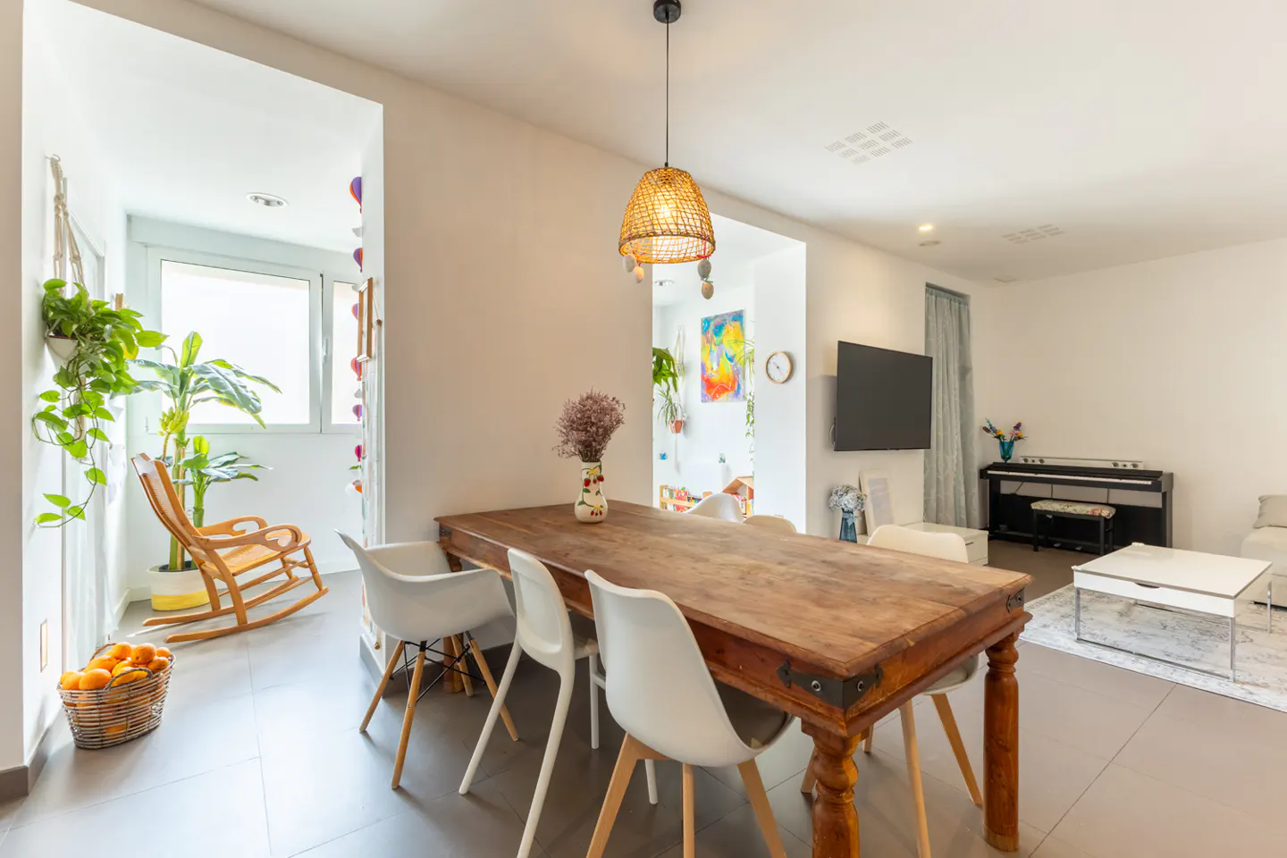 Bright, open-concept living space with a rustic wood dining table, white chairs, and a woven pendant light. A rocking chair and plants sit near a window.