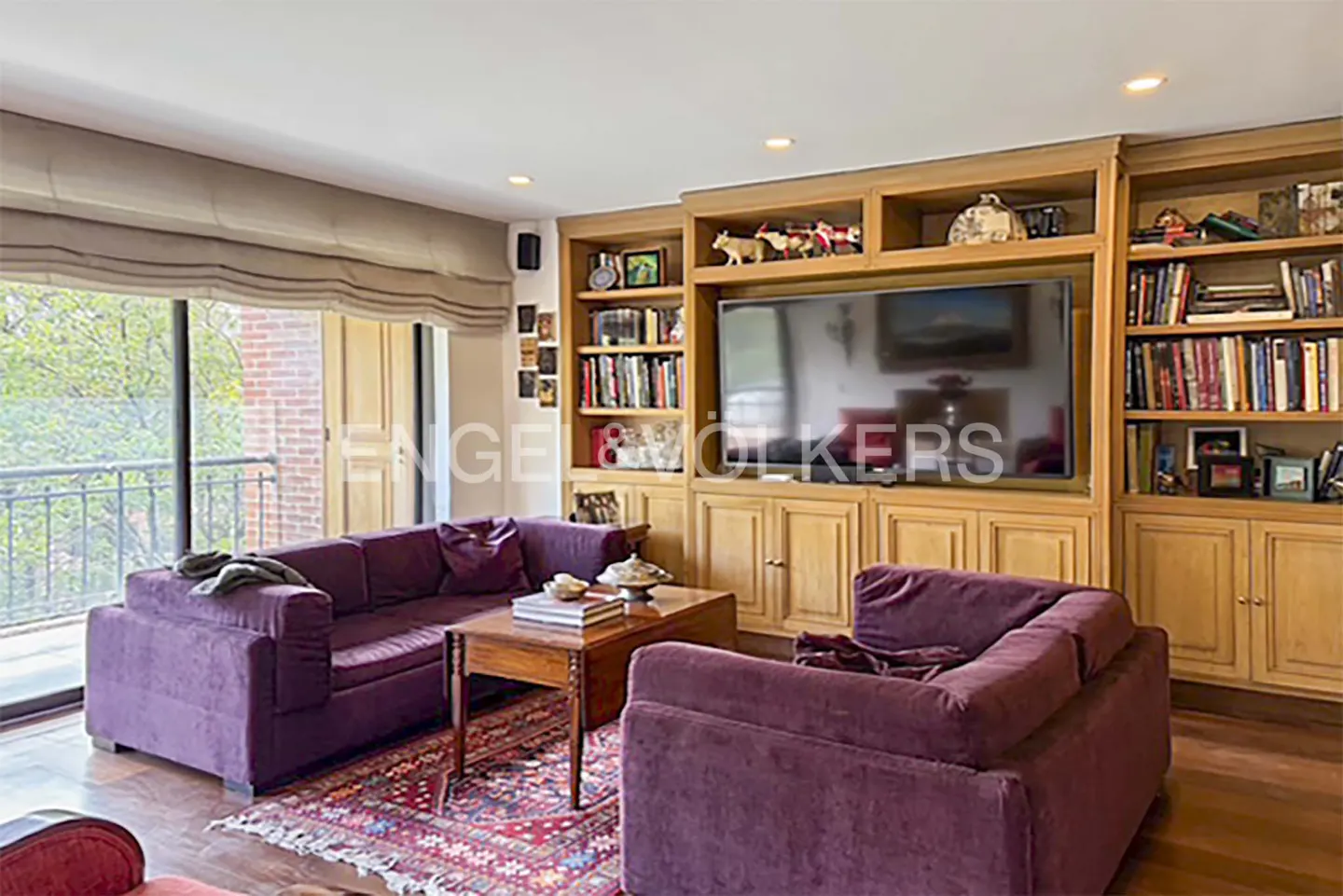 Living room with two purple sofas, a wooden coffee table, a patterned rug, and a large built-in wooden bookshelf with a TV.