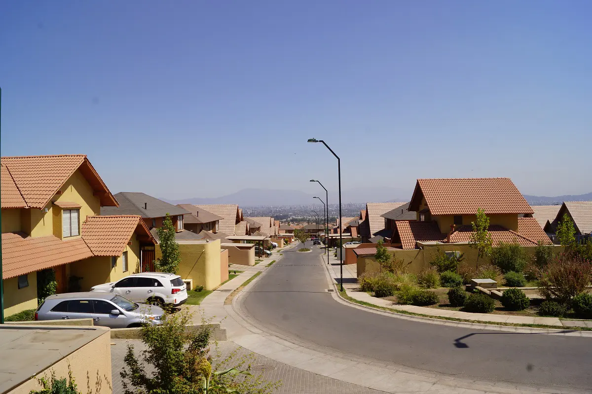 Residential street view with tan houses, orange tile roofs, and parked cars under a clear blue sky.
