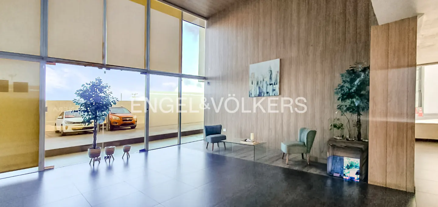 Modern lobby with floor-to-ceiling windows, gray tile floor, and wood-paneled walls. Two chairs and a glass table sit near an Engel & Völkers logo.