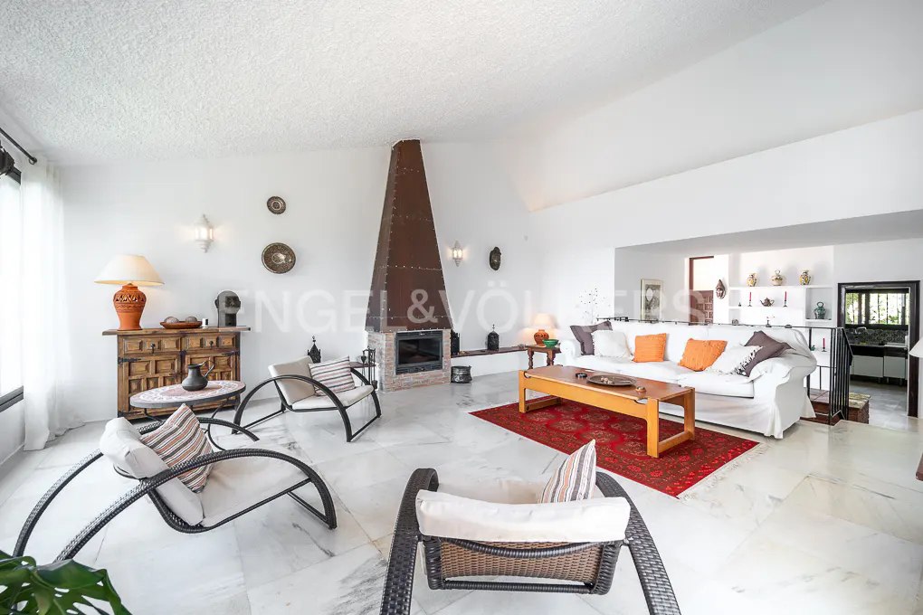 Bright living room with white walls, marble floors, and a tall brown fireplace. Wicker chairs and a white sofa with orange pillows add warmth.