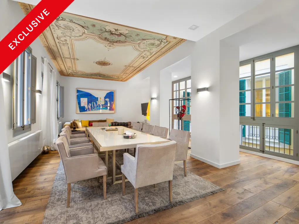 A bright dining room with a long table, beige chairs, and a decorative ceiling. Hardwood floors and large windows add to the room's elegance.
