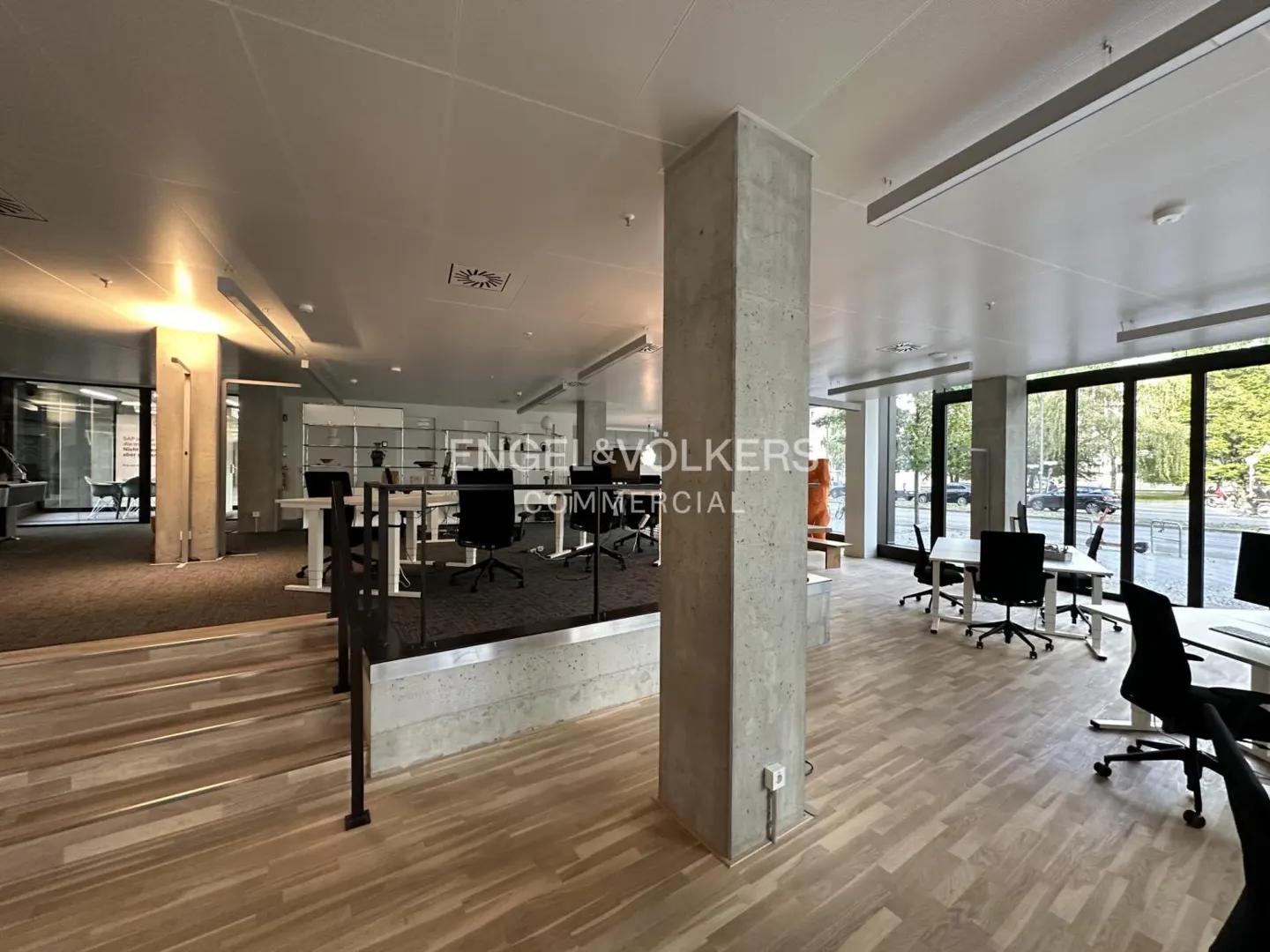 Bright office space with wood floors, concrete pillars, and black office chairs at white desks. Large windows offer natural light. Engel & Völkers logo visible.