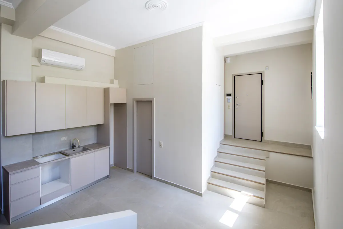 Minimalist apartment interior with beige cabinets, sink, and stairs leading to a door. The walls are painted in a light neutral tone.