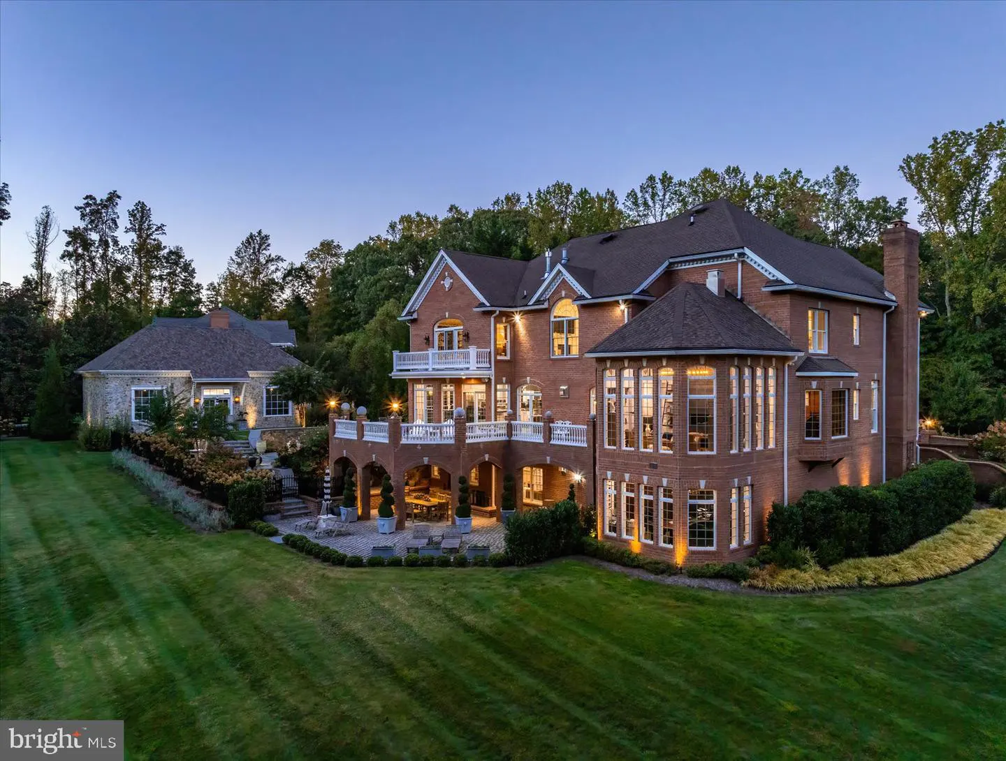 A large, two-story brick house with white balconies and a dark roof, surrounded by green lawn and trees.