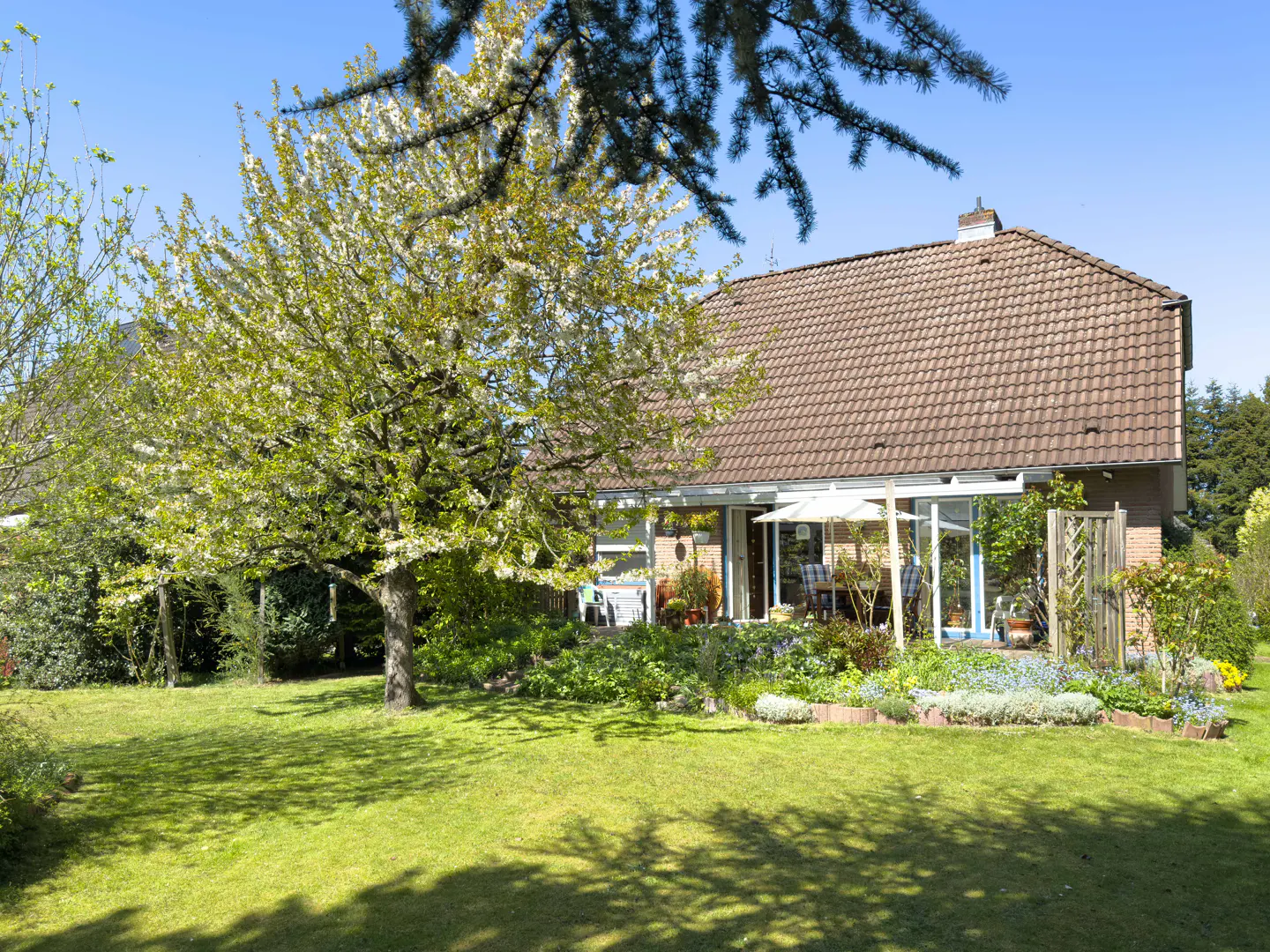 A brick house with a brown tiled roof sits behind a lush green lawn, a blooming tree, and a garden patio on a sunny day.