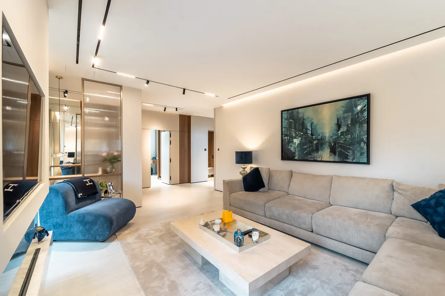 A modern living room with a beige sectional sofa, a light wood coffee table, and a blue accent chair. An abstract painting hangs on the wall.