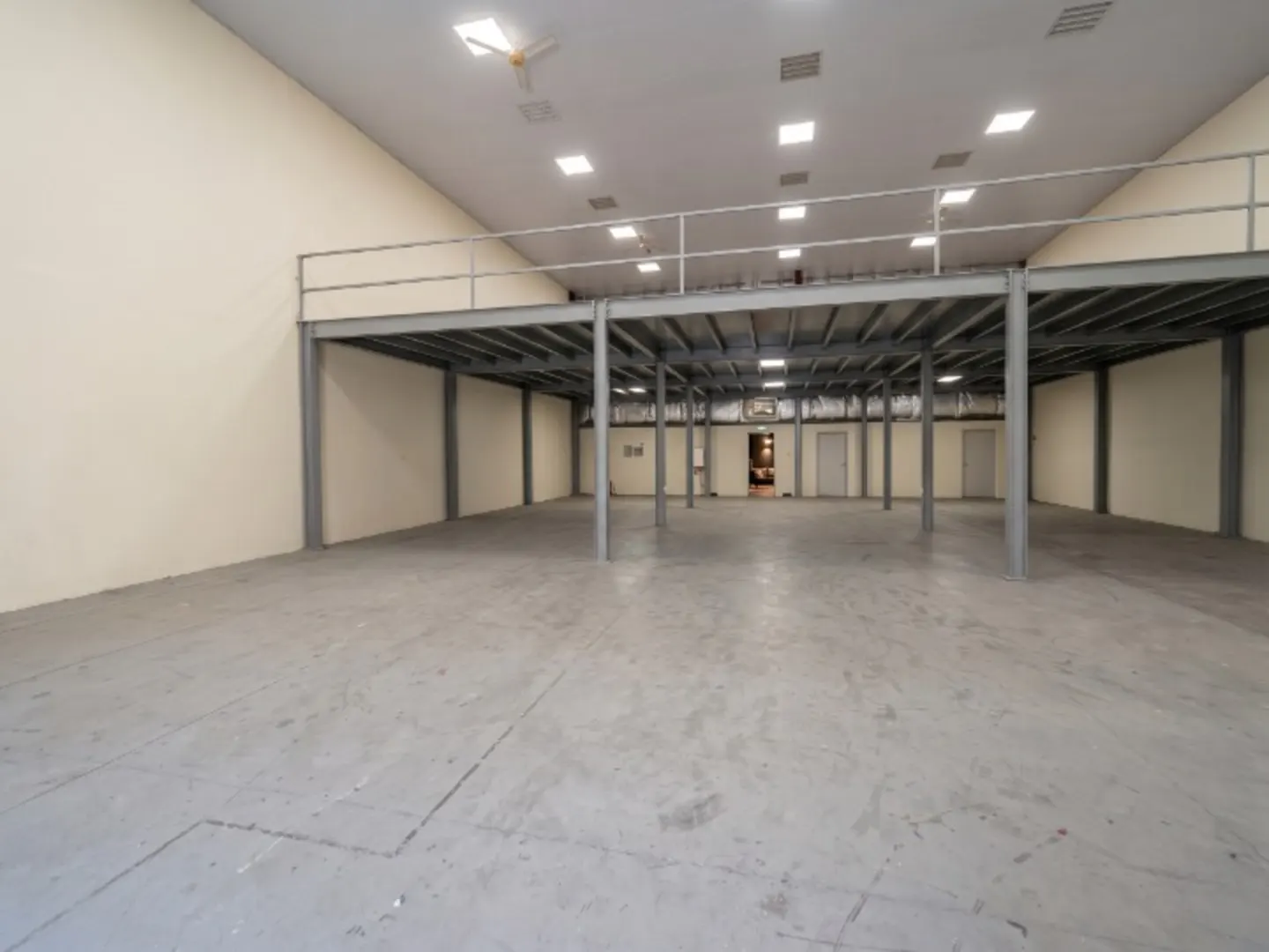 A large, empty warehouse with a gray concrete floor and a metal mezzanine. The walls are cream-colored, and the ceiling has fluorescent lights.