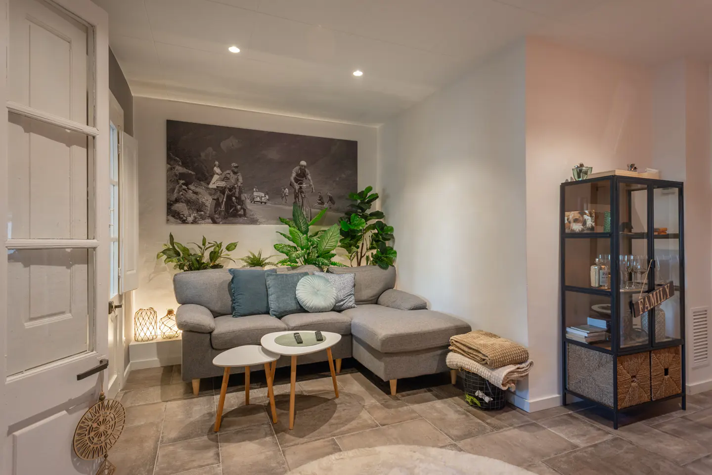 Living room with gray sectional sofa, blue pillows, two white tables, and a black and white cycling photo on the wall.