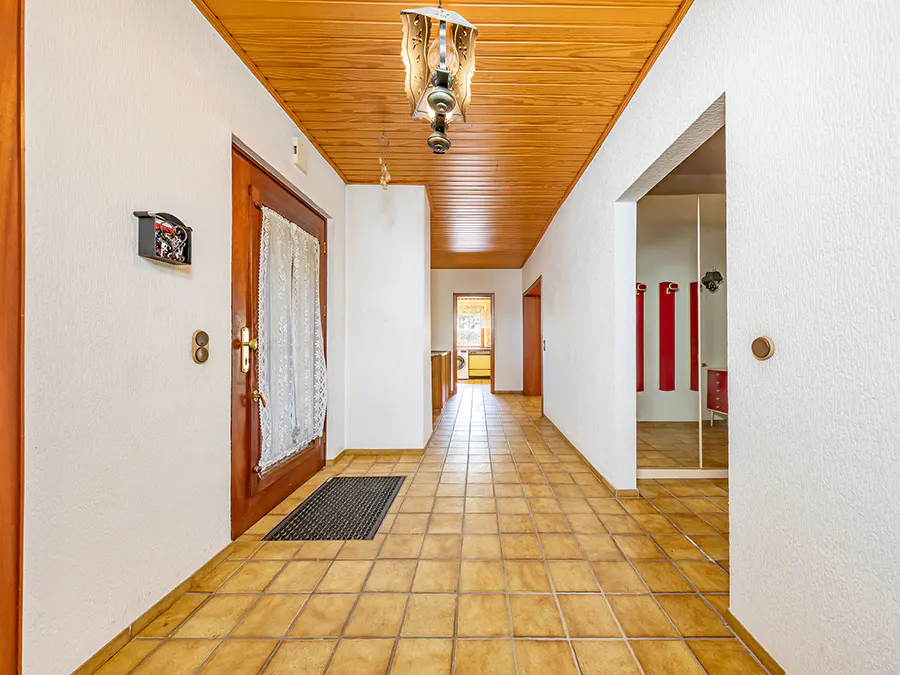 A hallway with tiled floors, wood-paneled ceiling, a decorative light fixture, and a wooden door with a lace curtain.