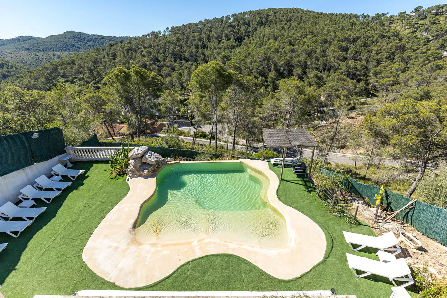 Aerial view of a kidney-shaped pool with turquoise water, surrounded by green turf, lounge chairs, and a forested hillside.