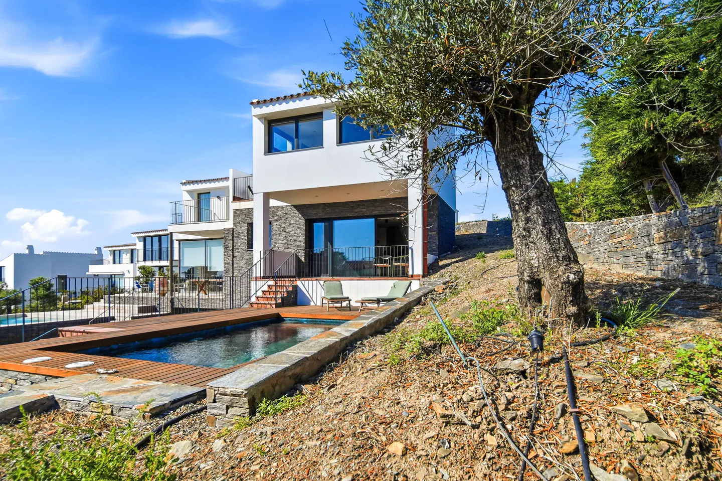 Modern white house with a pool and wooden deck. Lounge chairs sit near the pool. A tree is on the right, with a blue sky above.