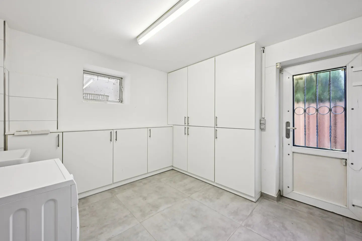 Bright laundry room with white cabinets, a washing machine, and a door with a decorative window. Gray tile floor and a small window.