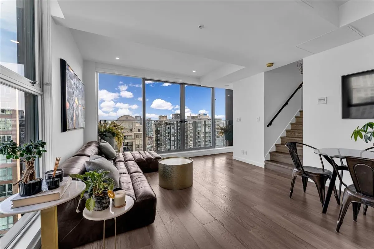 Bright living room with a brown leather couch, wood floors, and a city view through large windows. A staircase is visible in the background.