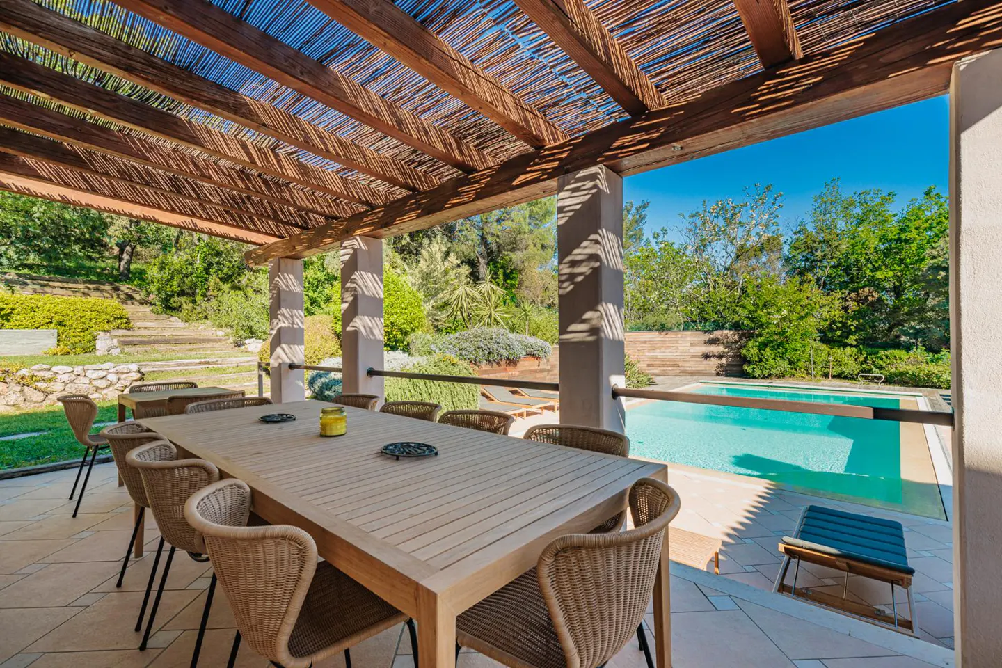 Outdoor dining area with a long wooden table and wicker chairs under a pergola, overlooking a pool and lush greenery.