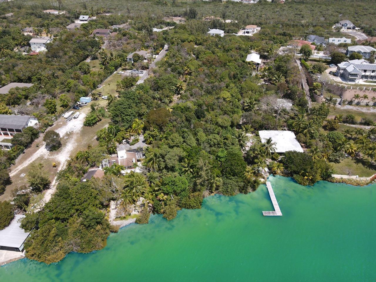 Aerial view of a waterfront property with lush green trees and turquoise water. A small dock extends into the water. Houses are scattered among the trees.