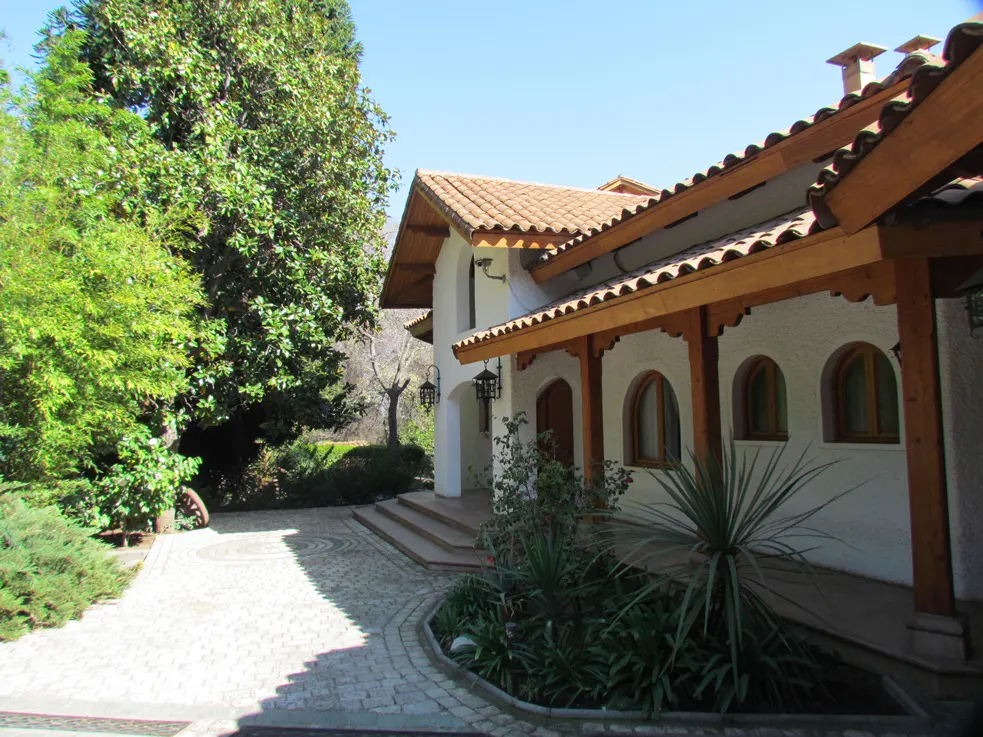 Exterior of a white stucco house with a red tile roof and wooden beams, surrounded by lush greenery. A stone path leads to the entrance.